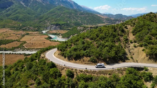 Aerial view of a campervan traveling, driving the iconic SH75 mountain road in Albania, winding through the rugged landscape above the vibrant turquoise waters of the Vjosa River in the summer sun.