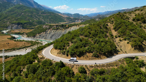 Aerial view of a campervan traveling, driving the iconic SH75 mountain road in Albania, winding through the rugged landscape above the vibrant turquoise waters of the Vjosa River in the summer sun.