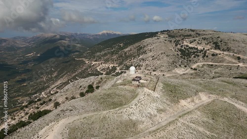 A white dome structure and building sit atop a rocky, green hillside. Winding dirt roads lead to the site, surrounded by mountains and a valley.
