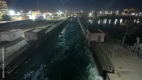 A night view of the Esna Lock in Luxor, Egypt, showing a ship navigating through the water channel with illuminated industrial structures.