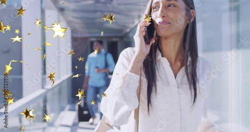 Walking woman in white blouse holding smartphone to ear in glass walkway, rolling suitcase behind