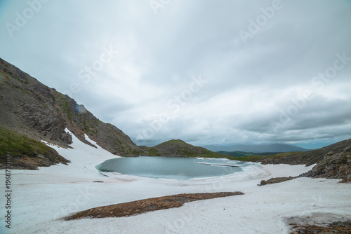Alpine lake among snows in hanging valley with view to green hills far away u...