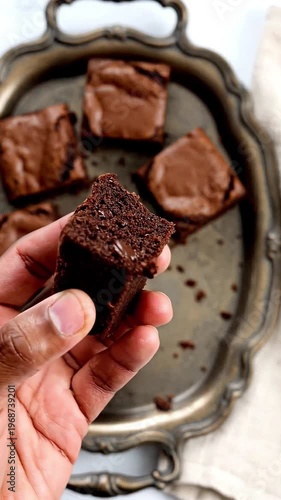 Hand holding a brownie piece with more brownies on a tray  
