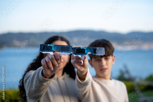 Teenagers showing solar eclipse glasses to camera outdoors