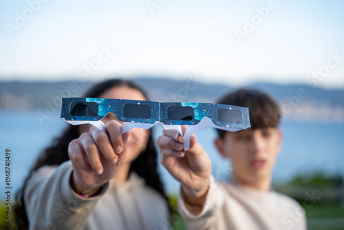 Teenagers showing solar eclipse glasses to camera outdoors