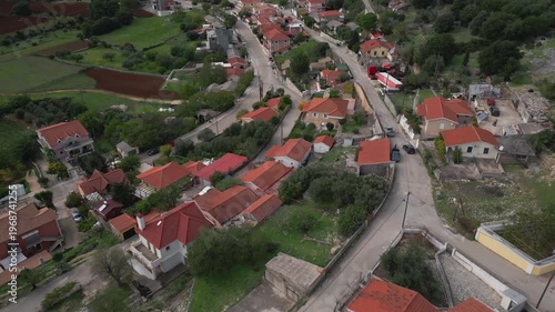 A village nestles among green fields and trees. Roads wind past houses with red tile roofs and some with modern facades.