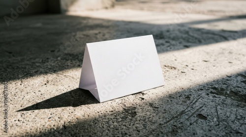 A blank white tent card stands on a concrete floor with dramatic diagonal light and shadow patterns, creating a minimalist and stark mood.