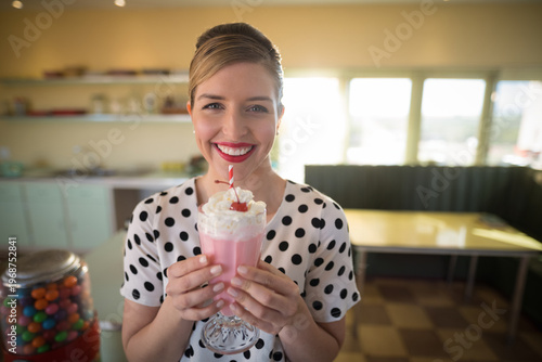 Pink milkshake sitting in footed glass on yellow diner table near window, gumball machine glowing