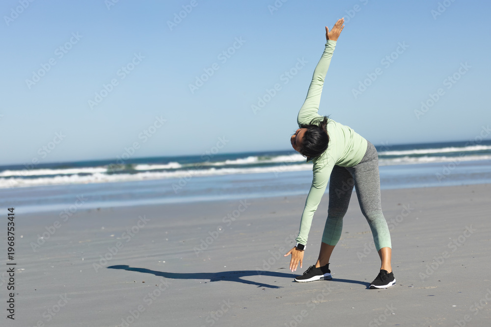 Fototapeta premium Mid adult Asian woman doing upright side-bend on beach with waves, tracker and sneakers, copy space