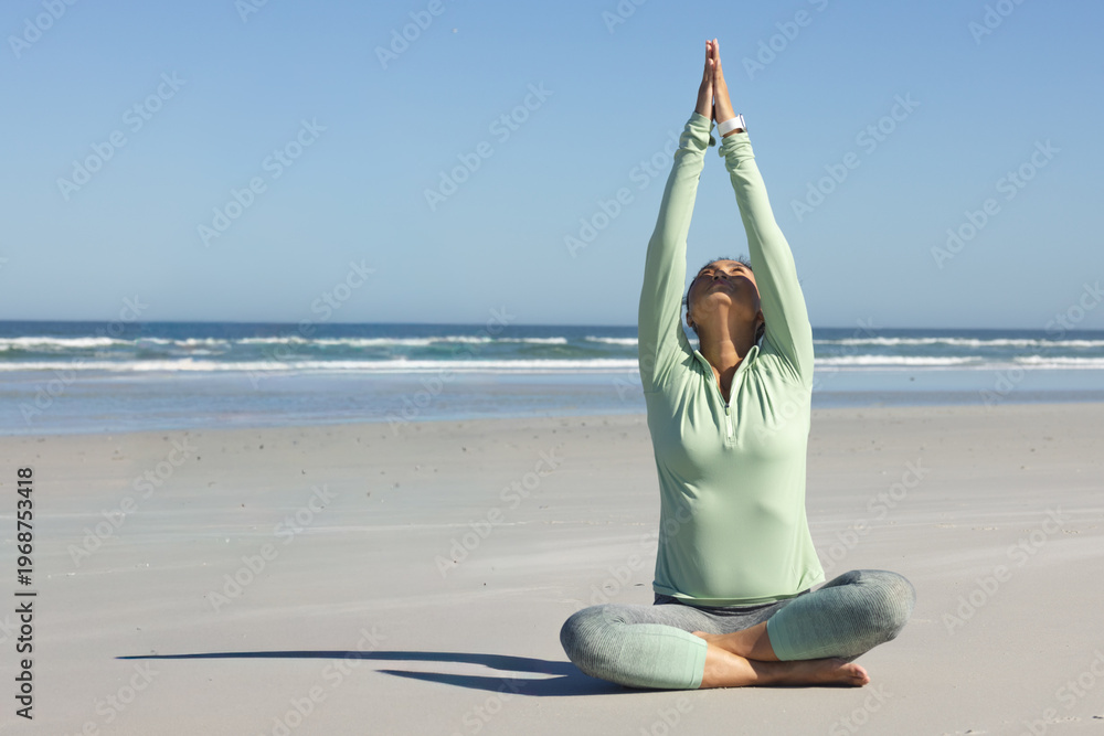Fototapeta premium Mid-adult Asian woman doing yoga, arms raised cross-legged on sand in athletic top, leggings, watch