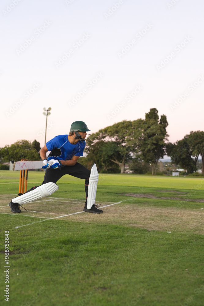 Obraz premium Adult male cricket batter holding wooden bat in helmet on pitch at dusk with stumps behind