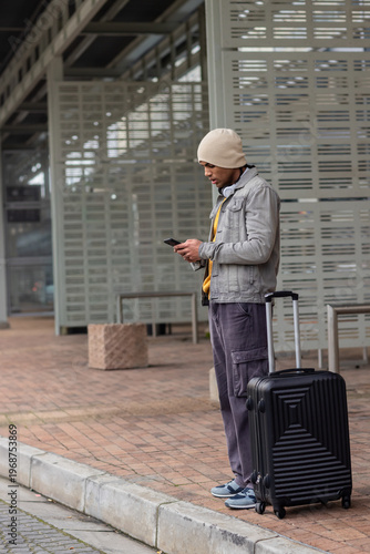 African American man standing at transit shelter checking phone in beanie with suitcase, copy space