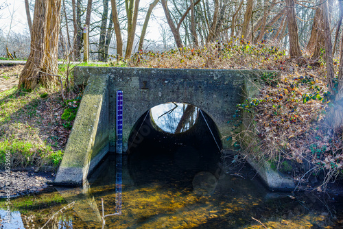 Pedestrian bridge with hydrometric scale and arch or vault through which water flows amidst wild vegetation and bare trees, low water levels reflected on surface, winter day in Maaseik, Belgium