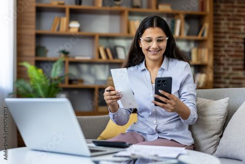 Canvas Print Young indian woman checking receipts on smartphone, doing calculations for taxes