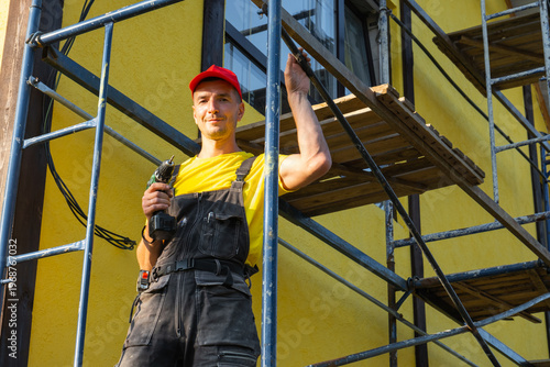 Builder with paint roller, scaffolding, ready for exterior wall works, home improvement, handyman instrument, service visit, professional construction repair.