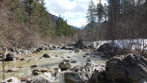 Aerial drone footage flying upstream above rocky mountain river through forest valley with remaining snow