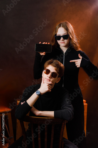 Cool teenagers posing in a dark studio, girl showing a black mobile phone screen and pointing at a sitting guy.