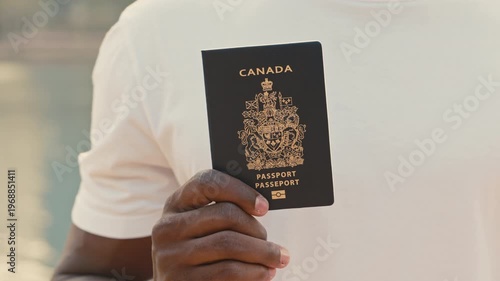 Black man in casual T-shirt holds passport of Canada. African American guy shows document of formal approval for permanent residency. Male celebrates newly granted citizenship