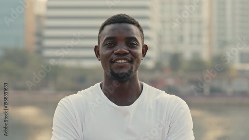 Portrait of cheerful African American man looking at camera while standing against urban buildings. Guy in casual T-shirt exudes happiness standing on city embankment