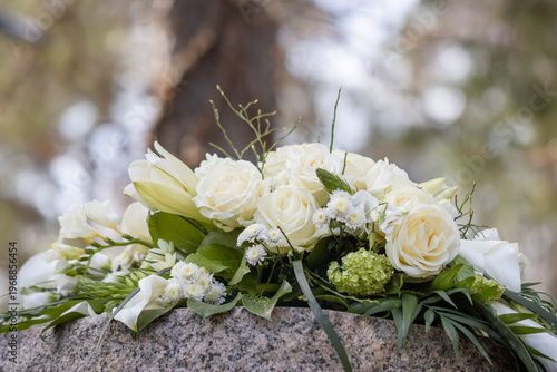 White funeral flowers on a memorial stone in a cemetery. A beautiful memorial bouquet of white roses and other white flowers in April. Empty space for text.