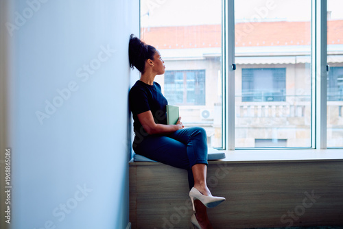 фотографія Woman sits on window ledge in modern office, holding a book and gazing out thoug