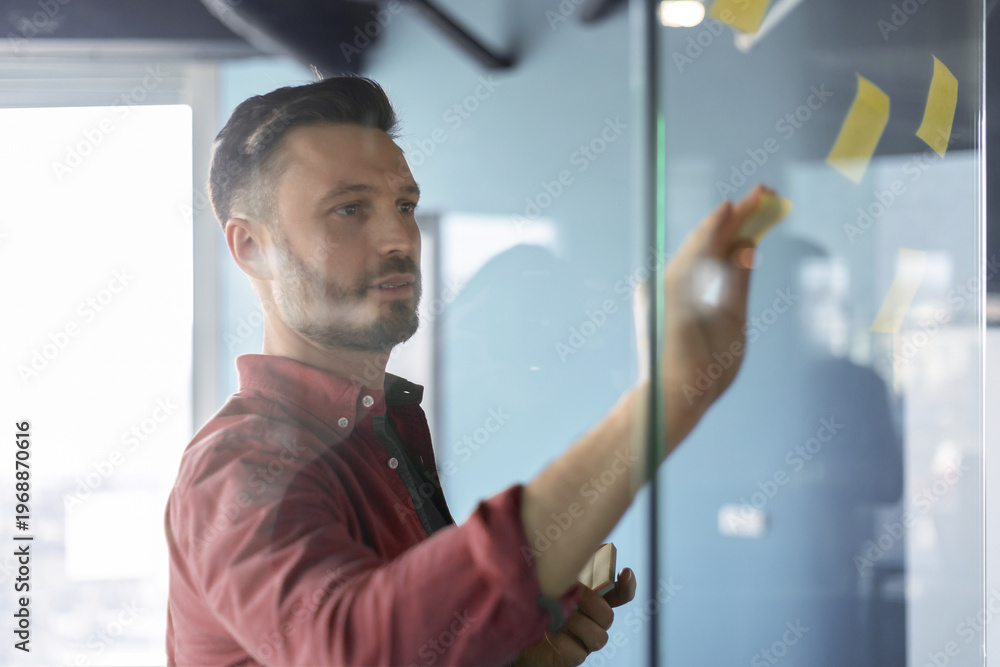 Naklejka premium A man is writing on a glass board in an office. He is focused on arranging sticky notes while collaborating with others. The setting shows a modern workspace with a view outside.