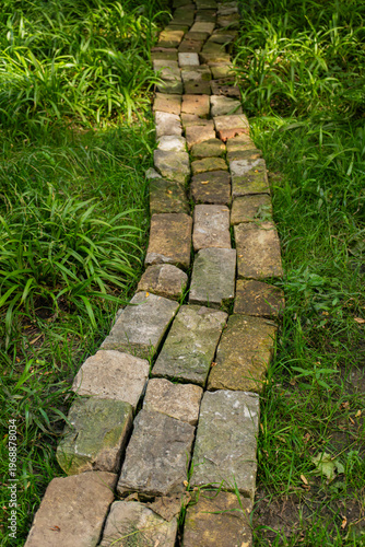 Принти на полотні Narrow straight path made of irregular old weathered stone and brick pavers running through lush green grass, top down perspective
