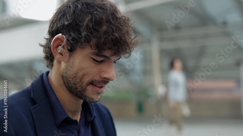 Portrait of curly haired Hispanic man manager with hearing aid stands on street. Latin corporate employee with disability smiles at office buildings. Diversity in workplace care