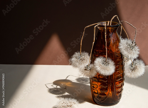 Quadro su tela Artistic still life with dried dandelion flowers in a vintage brown glass vase illuminated by dramatic sunlight