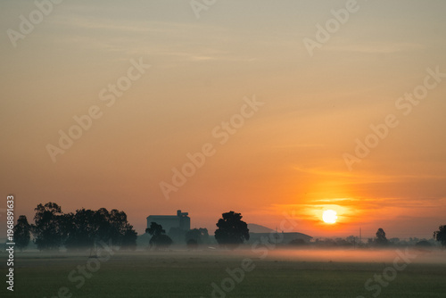 Sunrise over misty countryside with industrial silhouettes — a serene blend of nature and structure