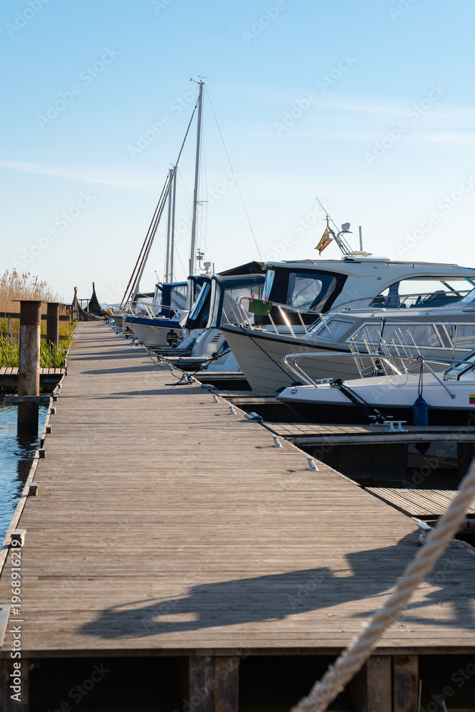 Fototapeta premium Sunny Marina Scene with Moored Boats and Wooden Pier Under a Clear Blue Sky, Inviting Serene Waterside Leisure