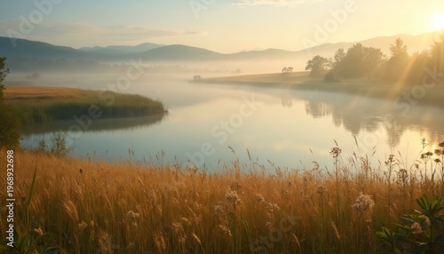 Golden sunrise beams through fog over calm lake and wheat fields. Mist blankets hills and trees creating serene landscape. Gentle water reflects soft light, peaceful dawn.