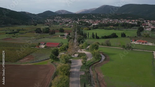 An aerial view captures a village nestled among rolling hills and verdant fields. A road winds through the landscape, leading to a cluster of houses with red-tiled roofs.