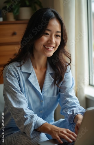 Young woman in blue striped pajamas works on laptop near window. She smiles gently. Natural morning light falls on her face, creating a cozy atmosphere at home. She wears comfortable clothes. Free Download Young woman in blue striped pajamas works on laptop near window. She smiles gently. Natural morning light falls on her face, creating a cozy atmosphere at home. She wears comfortable clothes. - GFXLoad
