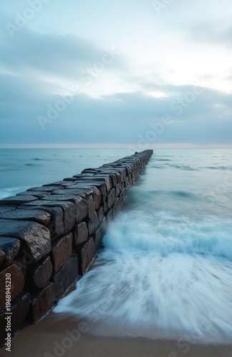 Stone breakwater extends into calm sea under cloudy sky. Water moves smoothly with long exposure effect showing wave motion against dark rocks and sand shore. Serene ocean backdrop.