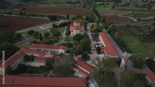 An aerial view of a monastery complex with a domed church, cloisters, and surrounding agricultural fields under a soft sky.