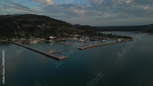 A serene marina with sailboats at dusk, nestled against a green, hilly coastline under a cloudy sky.