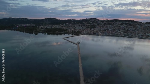 Sunrise paints the sky over a calm bay, reflecting a town nestled on a hill and a long pier reaching into the water.