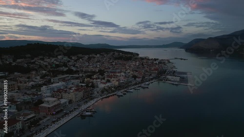 Aerial view of a coastal town at dusk. Buildings hug the shoreline, with boats docked in the harbor under a pastel sky.