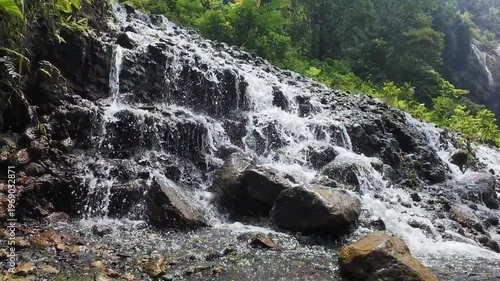 A beautiful natural landscape features a flowing mountain waterfall cascading over stones and green forest rocks into a summer stream