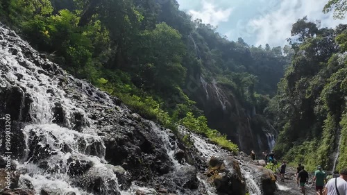 A beautiful natural landscape features a flowing waterfall in the Alps, where crystal water streams past green trees and rocks in a summer mountain forest valley