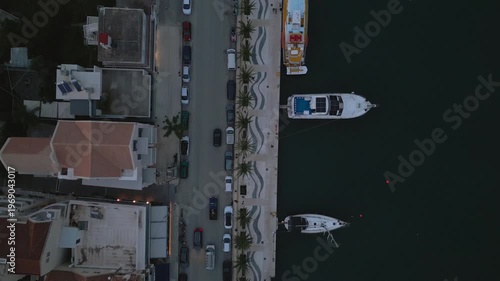 Aerial view of a coastal town with boats docked by a promenade and cars parked on a street.