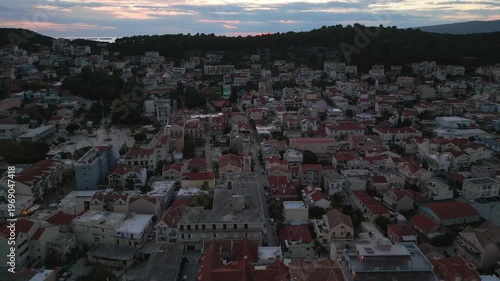 A picturesque coastal town unfolds at dusk, with a vibrant sunset painting the sky above terracotta rooftops and lush green hills.