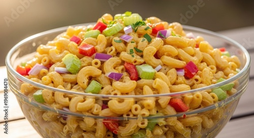 A bowl of macaroni salad with colorful vegetables in a clear glass bowl on a wooden table.