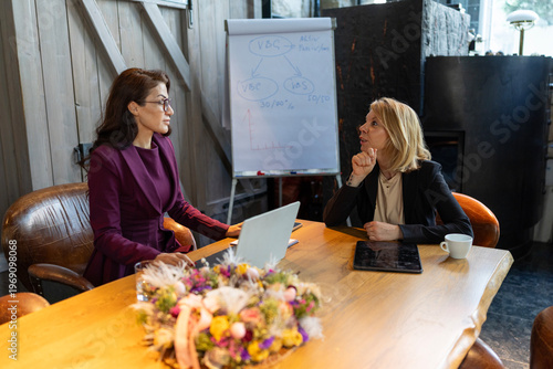 Two caucasian women business meeting in cozy industrial office, startup founder presenting strategy on laptop and flipchart, tablet and notebook on table, bouquet centerpiece, warm daylight,