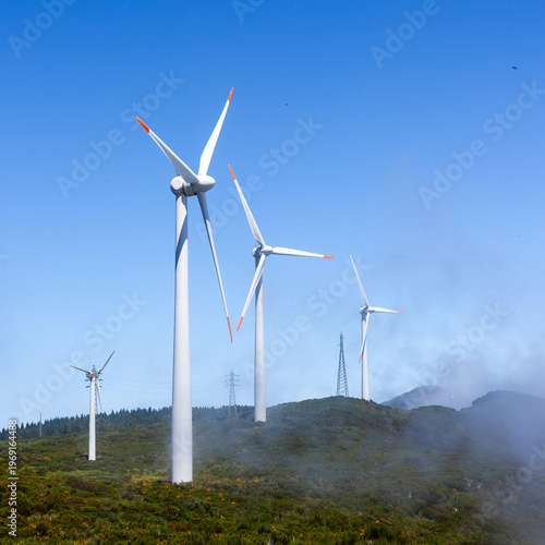 Wind turbines generating renewable energy on a hillside in Madeira. Green landscape with windmills under a clear blue sky. Power generators set against a mountainous backdrop.