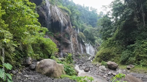 A beautiful summer landscape featuring a natural waterfall cascade flowing over rocks into a green mountain river stream deep within a lush forest canyon