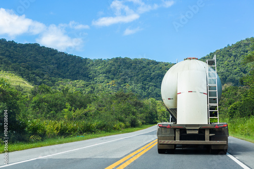 Fuel tanker truck driving on highway surrounded by lush green forest under blue sky, representing logistics and transportation in Brazil.