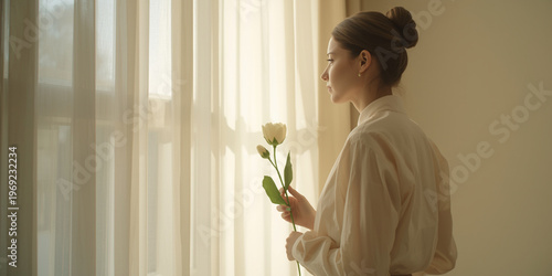 Serene young woman with elegant hair bun holding white tulip while gazing through soft backlit window curtains