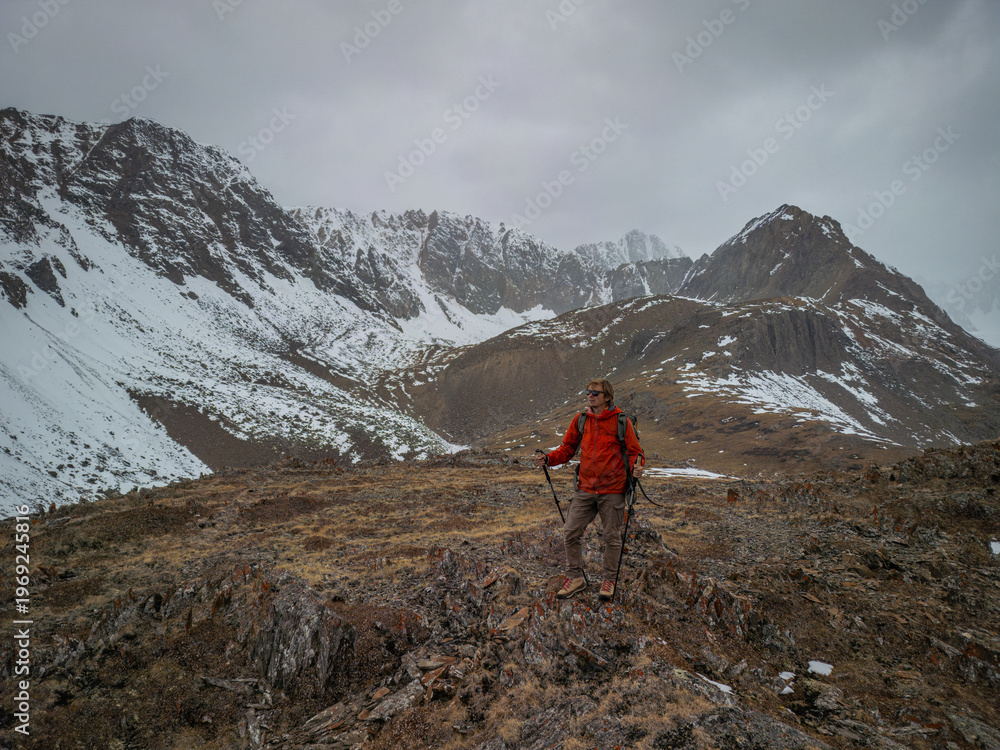 Obraz premium Male hiker in a red jacket with trekking poles standing on a rocky ridge against a backdrop of snowy mountain peaks under a cloudy sky, adventure travel and hiking concept.